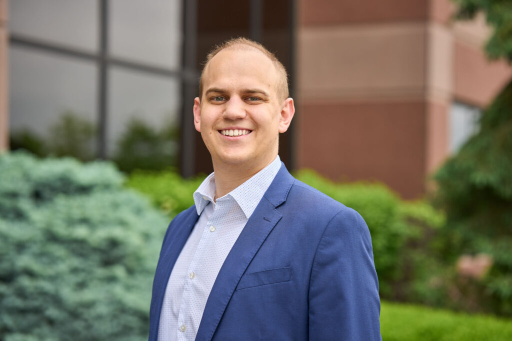 A man standing outside an office building wearing a navy suit, facing towards the left but smiling at the camera.